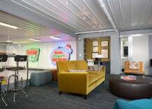 Man cave in garage features pub-height table lined with industrial barstools as well as mustard yellow velvet sofa paired with caramel colored Barcelona Chair and turquoise blue lounge chair across a round brown leather ottoman over gray carpeting.