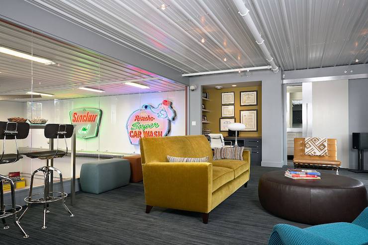 Man cave in garage features pub-height table lined with industrial barstools as well as mustard yellow velvet sofa paired with caramel colored Barcelona Chair and turquoise blue lounge chair across a round brown leather ottoman over gray carpeting.