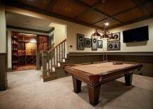 Fabulous basement games room with separate wine cellar. The basement features tongue and groove paneling in a khaki green with light yellow upper walls. The ceiling is coffered with dark stained beams and a dark brown center panel. Framed records and a flat screen tv are mounted on the walls behind the pool table. The traditional pool table is lit by a three light copper pendant. The wine cellar features hardwood floors and custom wine storage. The wine cellar is lit by an armillary style pendant.