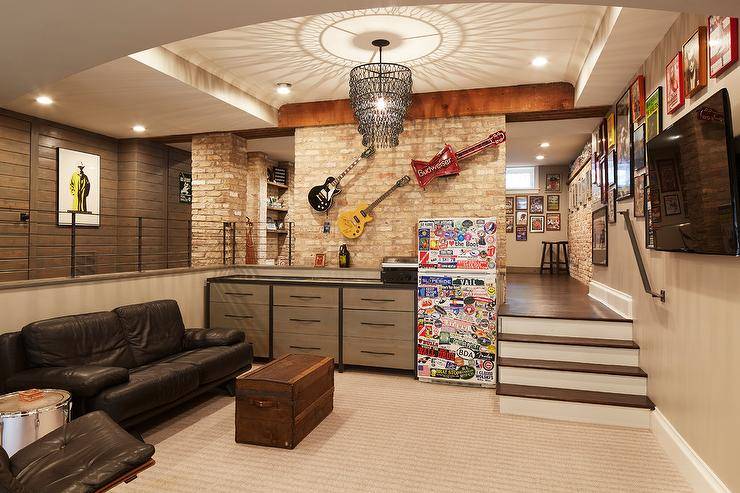 Well appointed sunken man cave features a black chain tiered chandelier illuminating a black leather sofa placed facing a trunk coffee table and a wall mount television mounted beneath framed art. This basement is also fitted with a sticker covered mini fridge placed beside a two toned black and gray dresser as decorative guitars are mounted above it on a brick wall.