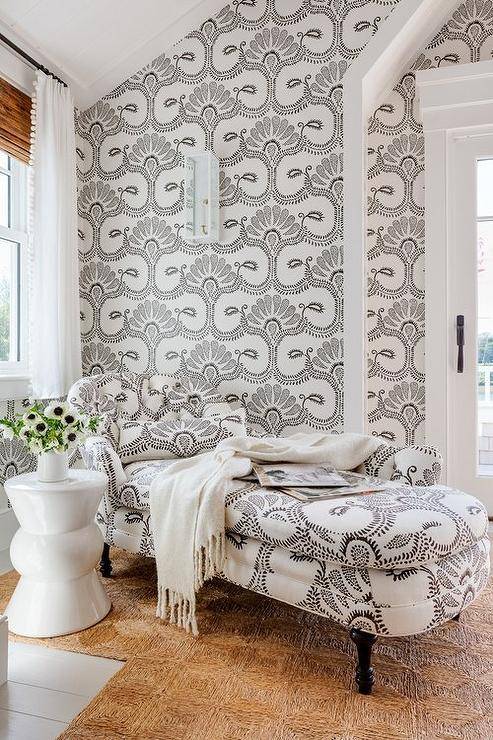 A black and white chaise lounge sit on a woven rug beside a white accent table and in front of walls clad in matching white and black wallpaper.