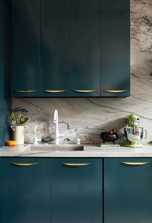 A modern white gooseneck faucet is mounted to a honed marble countertop over a gray sick fitted to glossy peacock blue flat front cabinets finished with curved brass pulls. Above the sick, peacock blue lacquered cabinets are mounted against a honed marble slab backsplash.