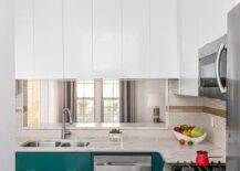 This well appointed contemporary kitchen is lit by a white modern chandelier fixed in front of a stainless steel oven range positioned between peacock blu cabinets topped with a calcutta quartz countertop and beneath a microwave surrounded by glossy white cabinets. A stainless steel dual sink with a polished nickel gooseneck faucet is located under a pass through framed by white penny tiles accented with gold grout and white feather tiles.