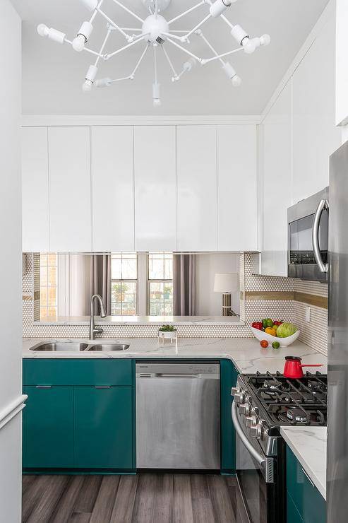 This well appointed contemporary kitchen is lit by a white modern chandelier fixed in front of a stainless steel oven range positioned between peacock blu cabinets topped with a calcutta quartz countertop and beneath a microwave surrounded by glossy white cabinets. A stainless steel dual sink with a polished nickel gooseneck faucet is located under a pass through framed by white penny tiles accented with gold grout and white feather tiles.