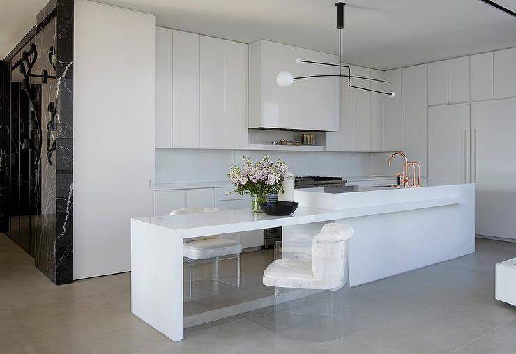 Spacious modern white kitchen features white velvet and acrylic stools at kitchen island dining table, an island sink with copper faucets and white flat front cabinets.