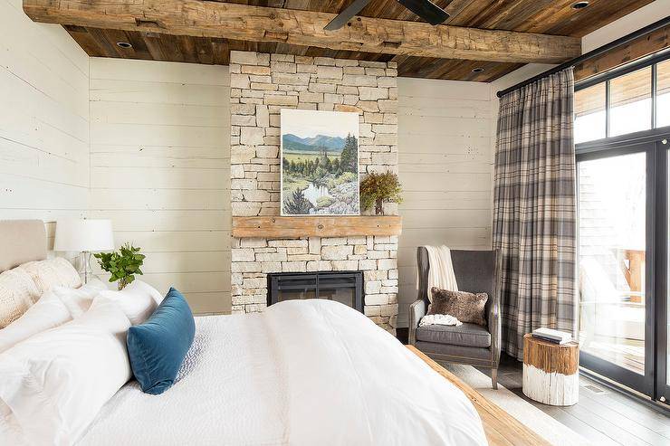 Country bedroom design with a rustic theme showcasing gray plaid curtains, a white bed, and a stone fireplace. A dip-dyed stool table next to a gray wingback chair adds a cozy vibe to a corner allowing for a sweet reading area. Rustic theme doesn't take away from a relaxing retreat design with decorative accents including a blue jewel velvet pillow, white throw blanket, wood mantle on fireplace and wall art on display. Rustic ceiling beams layered on planks adds a decorative and natural atmosphere.