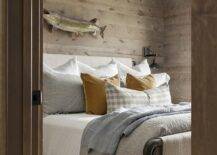 Rustic cabin bedroom features a fish mounted on plank walls over a vintage wood and metal bed accented with mustard yellow velvet pillows.