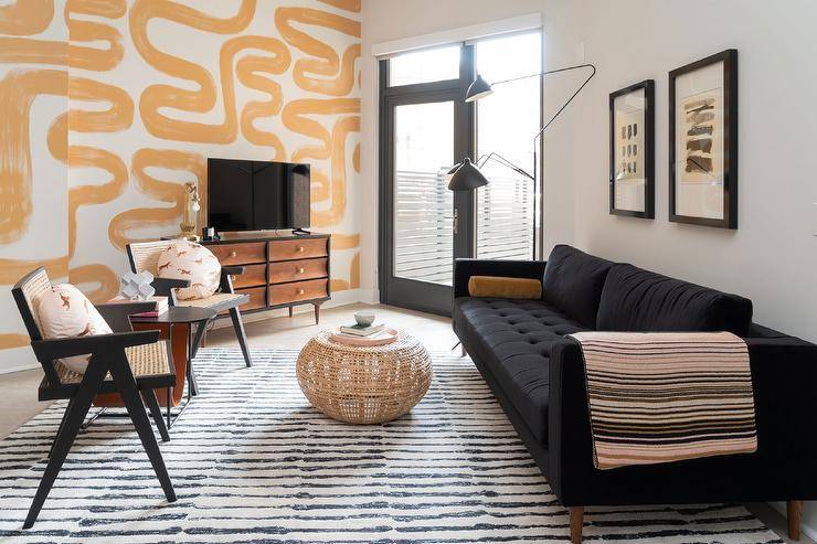 Contemporary black and yellow living room features art hung over a black velvet tufted sofa accented with mustard yellow velvet pillows. A wicker stool coffee table sits on a black and white striped rug in front of the sofa, while gold and black cane chairs are positioned facing the sofa. A flat panel TV sits on a mid-century modern TV cabinet in front of a mustard yellow painted accent wall. The room is lit by a matte black tripod floor lamp.