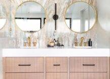 Modern bathroom features round brass mirrors mounted on a pink and gray marble slab accent wall over a beige reeded double washstand with brush gold faucets.