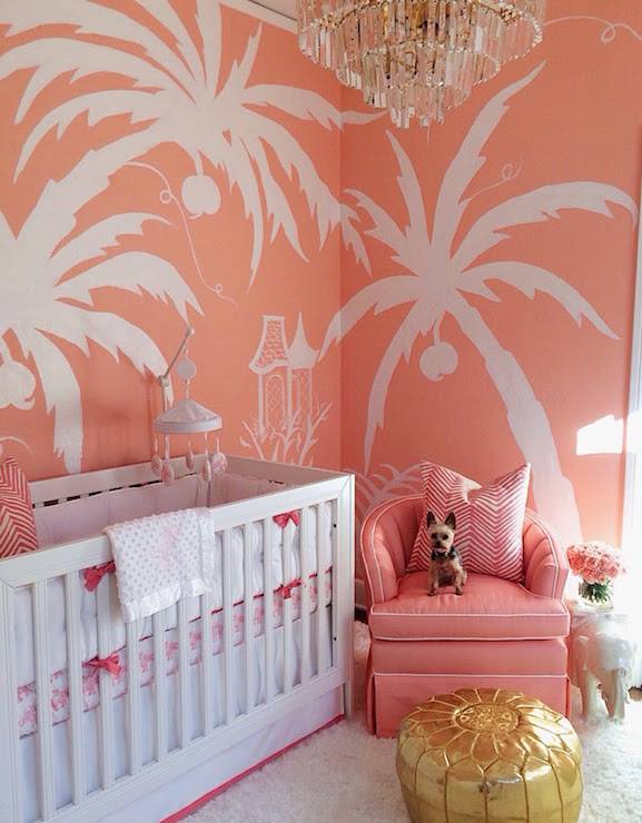 Coral pink nursery features walls painted coral, Sherwin Williams Jovial, adorned with a hand painted palm tree mural by artist EEK. A glass art deco chandelier illuminates a white crib dressed in white and pink elephant crib bedding, a Quadrille Petite Zig Zag Fabric pillow and a white and pink crib mobile next to a salmon pink chair and a Two's Company Elephant Side Table topped with pink roses as well as a gold Moroccan pouf atop a white sheepskin rug.