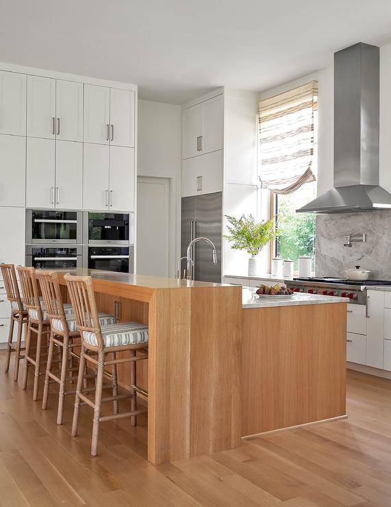 Kitchen features brown rattan stools with black striped cushions at a wooden waterfall breakfast bar and white cabinets with nickel pulls.