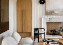 LIving room features white boucle low lounge chairs on a white and gray striped wool rug, an arched doorway, a white reeded accent wall and a marble fireplace.