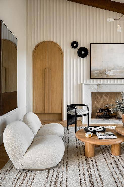 LIving room features white boucle low lounge chairs on a white and gray striped wool rug, an arched doorway, a white reeded accent wall and a marble fireplace.