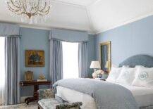 Sophisticated blue master bedroom features a blue velvet headboard that supports a bed dressed in a blue velvet pleated skirt, and white and blue monogrammed bedding, atop layered bedroom rugs. The bedroom boasts blue curtains and a gold Greek key mirror over a white gourd lamp. At the foot of the bed is a blue and white bench under a chandelier.