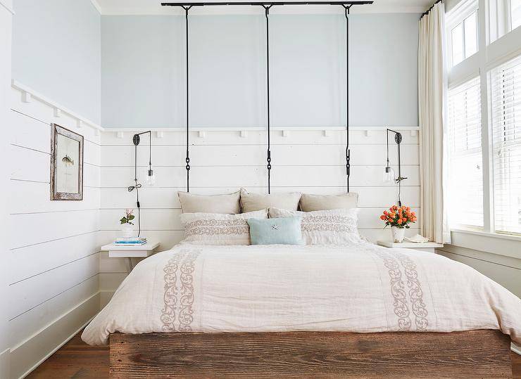 Romantic cottage bedroom features a hanging queen sized bed dressed in elegant cream colored bedding accented with matching shams and a blue pillow. The bed is flanked by white wall mount bedside tables illuminated by Edison lights fixed to a white shiplap trim lined with a light blue upper wall.