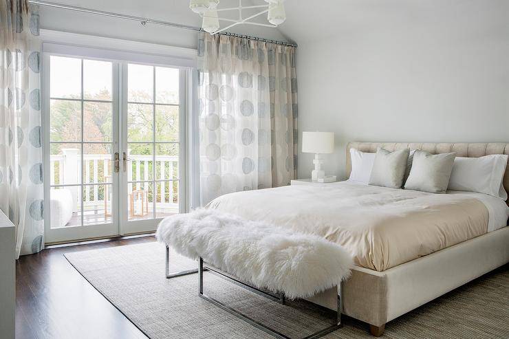 Romantic cream and gray bedroom boasts light gray walls and a cream suede bed accented with gold sheets and gray silk pillows. In front of the bed, a sheepskin bench sits on a gray bound sisal rug, while windows are covered in cream and gray dots curtains.