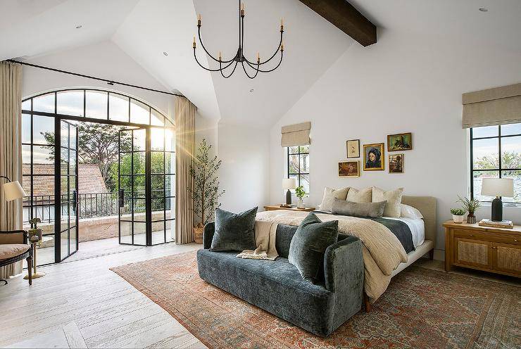 Romantic bedroom boasts a beige fabric platform bed dressed in beige and black bedding and placed on a red and tan vintage rug behind a black velvet loveseat, between cane cabinets, and under vintage-style gallery art. The cabinets are lit with black lamps and located under windows covered in beige roman shades. Beige curtains cover glass and iron French doors with sidelights.