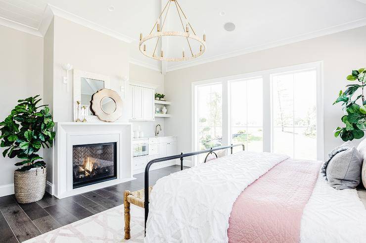 Master bedroom features a cream capiz mirror over a white beveled fireplace mantel, black bed with pink and white bedding, dark wooden floors, lit by a chandelier.