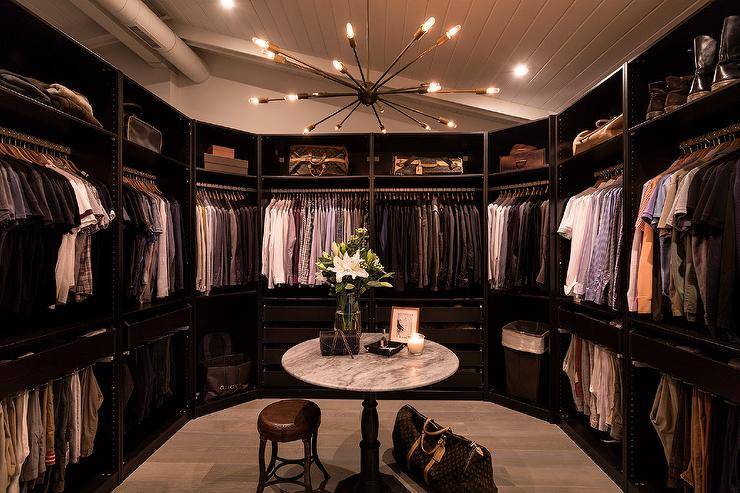 Chic men's large walk-in custom closet features Louis Vuitton luggage under a round marble accent table, illuminated by a sputnik chandelier.