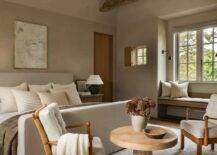 Restful cottage bedroom boasts rustic wood ceiling beams mounted to a gray vaulted ceiling over a natural linen slipcovered bed complemented with beige striped pillows. The bed is positioned against a gray wall under a rustic art piece. A round wooden table is flanked by brown wood chairs placed at the foot of the bed on a gray rug.