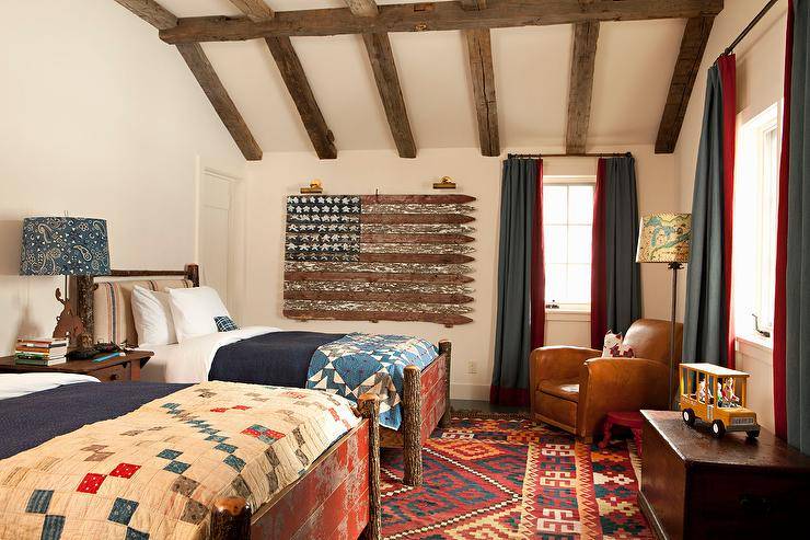 Country style bedroom features rustic tree branch poster beds with burlap headboards and farmhouse quilts, red and gray curtains, a corner leather accent chair lit by a floor lamp, a red print rug and wooden ceiling beams.