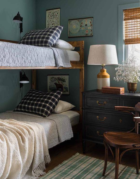 Shared boys' bedroom features a rustic bunk bed dressed in black and white bedding complemented with black plaid pillows. Black swing arm sconces light each bed, as art hangs from blue walls. A white and mustard yellow lamp sits on a black dresser finished with brass hardware and positioned beneath a window dressed in a bamboo roman shade. A wooden chair sits on a green plaid rug.