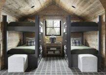 Attic boy's bunk bedroom features charcoal gray bunk beds with olive green bedding, a shared wooden nightstand lit by a gray lamp, an oak plank ceiling with oak plank walls, cube stools at the foot of beds and a charcoal gray rug.