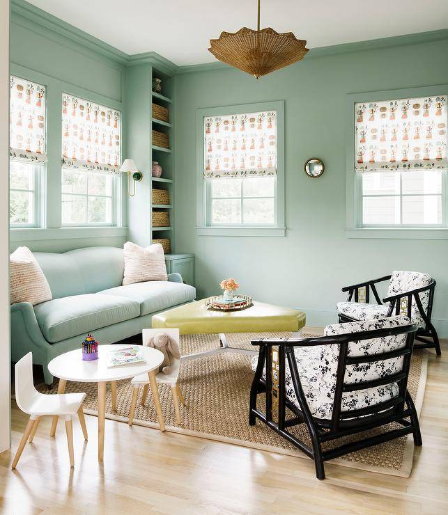 Welcoming living room boasts mint green walls framing windows located between mint green built-in shelves and over a mint green velvet sofa. The sofa sits on a tan bound sisal rug at a citron green leather triangle ottoman and is complemented with black chinoiserie chairs. Two white molded plastic kids' chairs sit at a round white kids' table. The room is illuminated by a rattan chandelier.