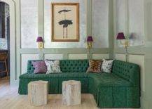 Eclectic living room features an emerald green tufted French sectional on sage green wall trim lit by scones with purple shades and dual hexagon accent tables.