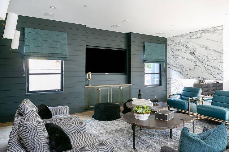 Turquoise blue roman shades hang from windows framed by hunter green shiplap and flank an inset TV fixed above turquoise blue built-in cabinets lined with gold trim. Side-by-side black and white chevron accent chairs and turquoise blue leather and brass chairs sit on either side of industrial nesting coffee tables placed on a gray rug.