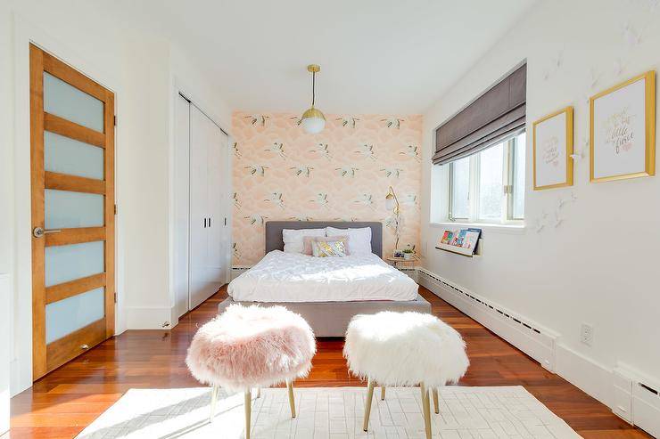 Lovely pink and gray teen girl's bedroom is furnished with pink and white faux fur stools placed on a gray rug in front of a gray upholstered bed dressed in white pintuck bedding lit by a white and gold light pendant. The bed is positioned against a wall clad in Harlequin Cranes In Flight Wallpaper and between white closet doors and a window covered with a gray roman shade hung over a white book ledge. Gold framed art hangs from a wall facing a wood door finished with frosted glass panels.