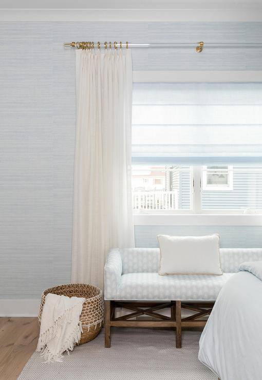 Light blue grasslcoth wallpaper frames a bedroom window dressed in ivory curtains hung from an acrylic and brass rod over a white and silver block print bench.