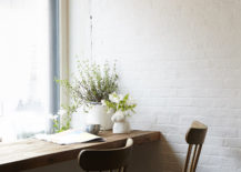 A wooden table is mounted on the wall by a large window, accompanied by two chairs. Above, a fan and lights hang.