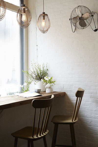 A wooden table is mounted on the wall by a large window, accompanied by two chairs. Above, a fan and lights hang.