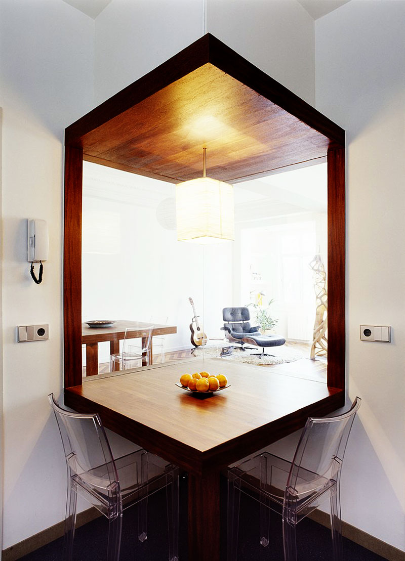 A wooden table with a colorful bowl of fresh fruit in the center is surrounded by two transparent chairs.