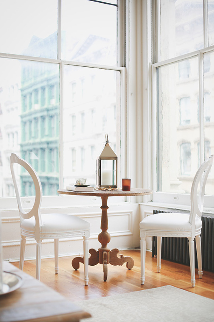 A room with large windows features a wooden table and chairs. The chairs have soft cushions and are positioned to enjoy the view outside.