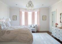 A crystal chandelier illuminates this elegant gray and pink girl's bedroom featuring a white bed placed on a gray rug and dressed in gray ruched bedding accented with pink shams. The bed faces a gorgeous gray French dresser positioned beneath a gray French mirror. Flanked by framed art, a bay window is covered with with pink curtains matching a pink cornice box and hanging behind a gray French chair.