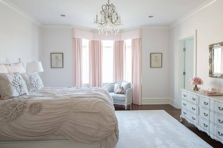 A crystal chandelier illuminates this elegant gray and pink girl's bedroom featuring a white bed placed on a gray rug and dressed in gray ruched bedding accented with pink shams. The bed faces a gorgeous gray French dresser positioned beneath a gray French mirror. Flanked by framed art, a bay window is covered with with pink curtains matching a pink cornice box and hanging behind a gray French chair.