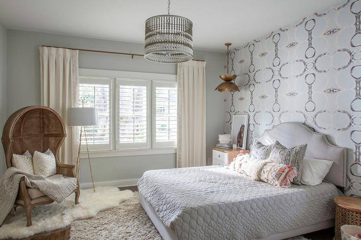 A white and gray beaded drum light pendant illuminates an elegant girl's bedroom featuring a gray arch headboard supporting a gray upholstered bed dressed in gray quilted bedding topped with silver pillows layered behind a Moroccan wedding black lumbar pillow. The bed is flanked by a wood stool table and a Bungalow 5 Berkeley 3 Drawer Side Table lit by a gold lotus chandelier hung in front of a gray and black abstract wallpapered accent wall. The bed sits on a gray wool rug, while a French cane dome chair topped with gold confetti pillows sits on a white sheepskin rug in a corner lit by a gold faux bois tripod floor lamp placed in front of a window dressed in white curtains complementing gray wall paint.