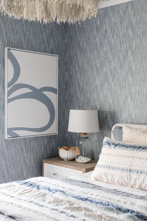 A glass bottle lamp sits atop a two-toned nightstand placed in front of a wall clad in blue textured wallpaper and beside a white slipcovered bed accented with white and blue bedding.