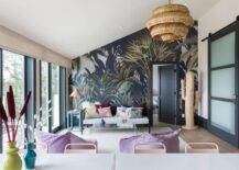 Living room features a black cane daybed with a blue bone inlay end table against a black and green accent wall, a clear acrylic waterfall coffee table atop a cream rug and a large woven cactus, illuminated by a woven chandelier.