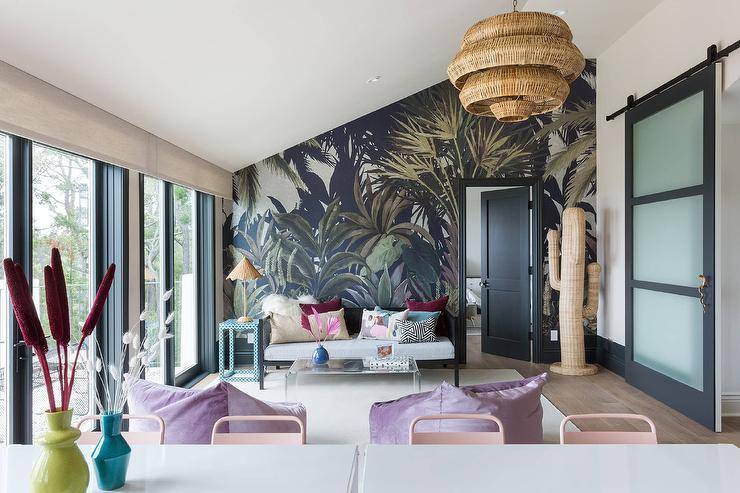 Living room features a black cane daybed with a blue bone inlay end table against a black and green accent wall, a clear acrylic waterfall coffee table atop a cream rug and a large woven cactus, illuminated by a woven chandelier.