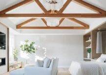 A glass icicles chandelier hangs from a truss ceiling in an elegant master bedroom boasting white his and hers loungers placed on a light gray rug facing a fireplace finished with a TV.