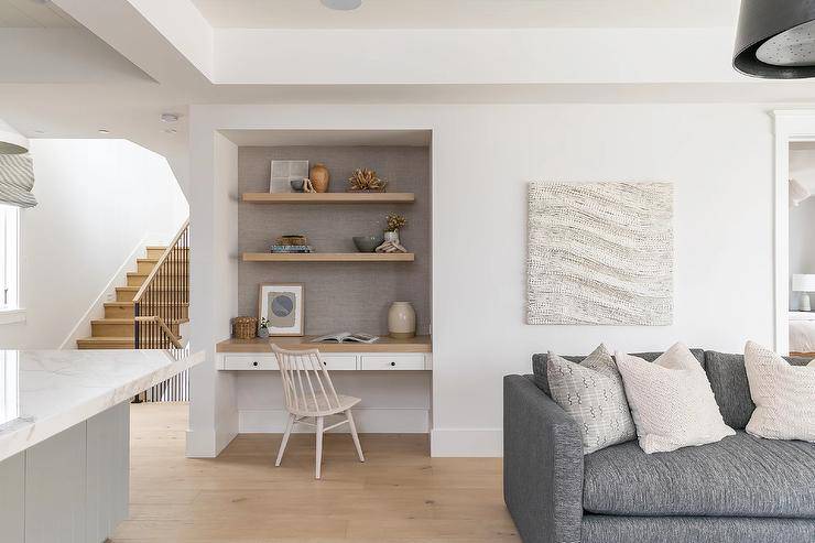 Wood floating shelves are mounted against taupe wallpaper and over a two-tone built-in desk paired with an off-white spindle chair.