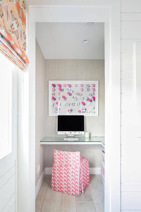 A pink stool sits at an l-shaped bench boasting a glass top and mounted under an aerial beach art piece hung from a gray wallpapered wall.