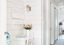 A blue stripe rug lines a cottage hallway boasting a white built-in desk matched with a white bamboo desk chair and positioned beside a white built-in bench with turquoise blue ikat cushions. The desk is fixed beneath stacked art mounted to a white tongue and groove wall beneath white overhead cubbies and lit by a cage sconce.