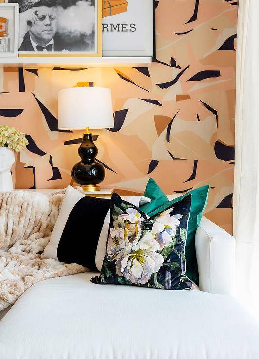 A white couch accented with black striped pillows is positioned beneath a floating picture shelf mounted to a wall covered in pink and black geometric wallpaper over a white and gold sofa table lit by black double gourd lamps.