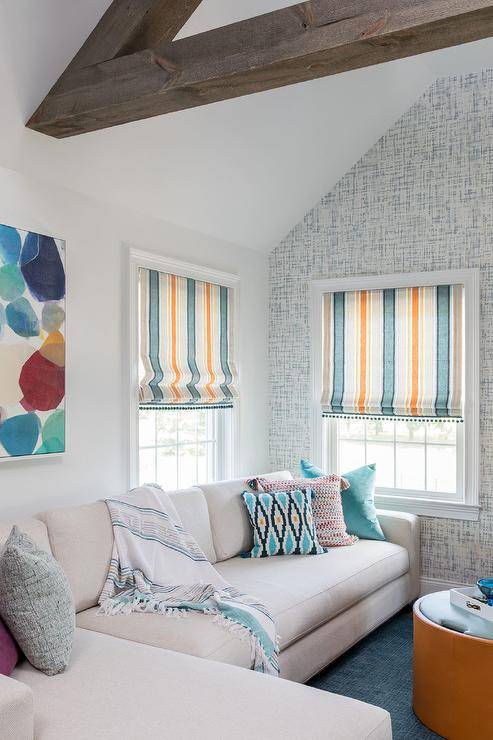 Beneath an abstract art piece and a window covered in an orange and blue striped roman shade, a light gray sectional is finished with purple, gray, and blue pillows. The sectional sits on blue carpeting facing a round orange and blue ottoman coffee table, while an accent wall is clad in gray and blue wallpaper.