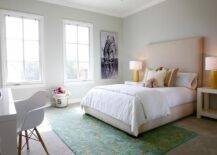 Restful teen girl's bedroom boasts gray carpet covered in a green and gold rug positioned beneath a tan upholstered bed accented with white hotel bedding topped with tan pillows and flanked by West Elm Parsons Side Tables lit by yellow faux bois lamps. A zebra print is hung from a light gray wall beside two side-by-side windows as an Eames Molded Plastic Arm Chair sits in front of the bed facing a white lacquered desk.