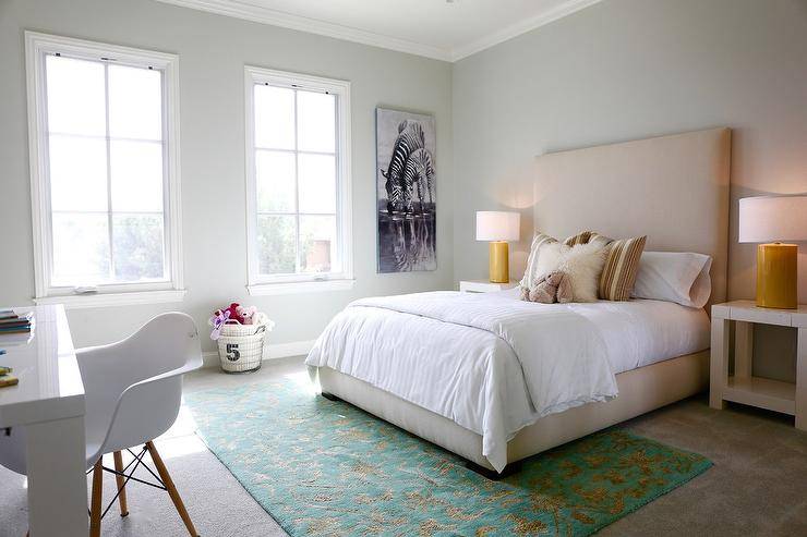 Restful teen girl's bedroom boasts gray carpet covered in a green and gold rug positioned beneath a tan upholstered bed accented with white hotel bedding topped with tan pillows and flanked by West Elm Parsons Side Tables lit by yellow faux bois lamps. A zebra print is hung from a light gray wall beside two side-by-side windows as an Eames Molded Plastic Arm Chair sits in front of the bed facing a white lacquered desk.